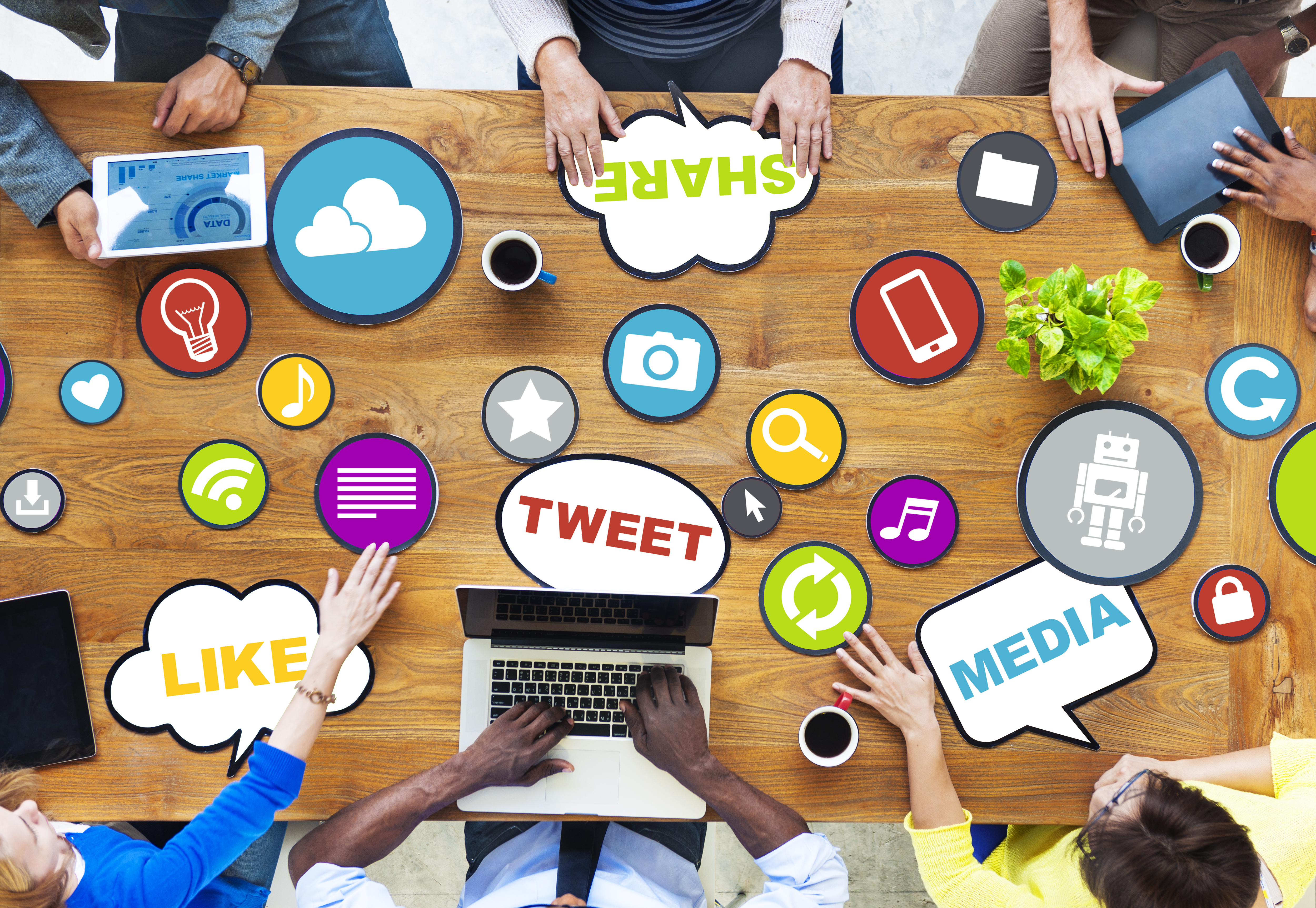 An overhead veiw of a group of people at a table on their laptops and tablets with 
            colorful animation around them and the words Media, Like, Tweet, & Share displayed